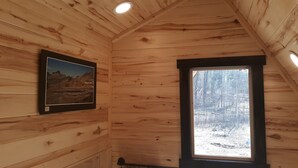 Interior de una cabaña de madera con una fotografía enmarcada en la pared, una ventana que da a un bosque nevado e iluminación empotrada en el techo.