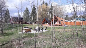 En rustik hytte med terrasse og udendørs siddepladser, omgivet af træer og en græsplæne.