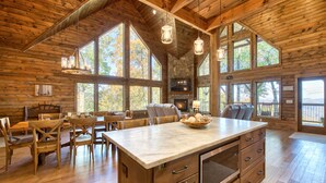 A spacious wooden cabin with a large dining area, a kitchen with a stone fireplace, and large windows offering views of the surrounding forest. - Newly Built 2021!! 4 BD 4.5 BA, Luxury Home, Mtn Views, Hot-tub! (Maggie Valley)