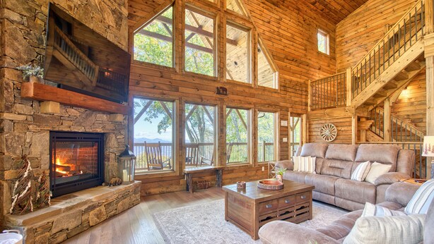 A rustic cabin interior with a stone fireplace, wooden floors, a large sofa, and a staircase leading to an upper level. - Newly Built 2021!! 4 BD 4.5 BA, Luxury Home, Mtn Views, Hot-tub! (Maggie Valley)