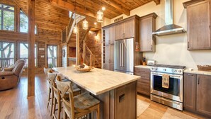 A spacious kitchen with wooden beams, a central island with bar stools, stainless steel appliances, and a view through large windows. - Newly Built 2021!! 4 BD 4.5 BA, Luxury Home, Mtn Views, Hot-tub! (Maggie Valley)