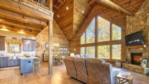 A spacious living area with a stone fireplace, wooden ceiling beams, and large windows offering a view of autumn foliage. - Newly Built 2021!! 4 BD 4.5 BA, Luxury Home, Mtn Views, Hot-tub! (Maggie Valley)