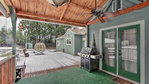 Un patio couvert avec un barbecue et un coin salon, une terrasse en bois et une maison avec des portes vertes.