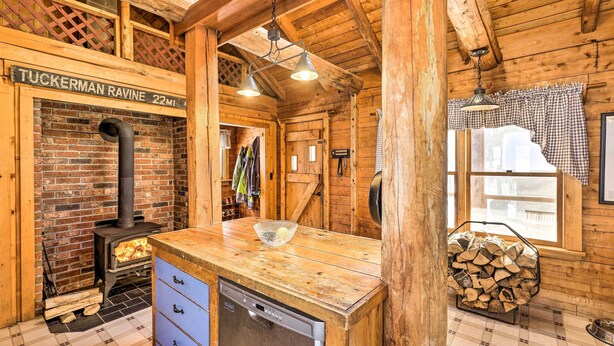 Ein rustikales Blockhaus-Interieur mit Kaminofen, Esstisch aus Holz und Blick ins Freie durch ein Fenster.
