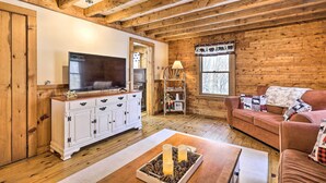 Ein gemütliches Blockhaus-Interieur mit Holzbalkendecke, einer weißen Unterhaltungsecke mit Fernseher, einem braunen Sofa mit Zierkissen und einem Holztisch mit Kerzen.