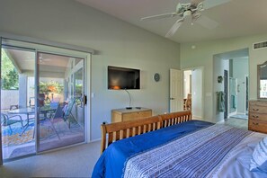 Une chambre avec un lit, un téléviseur fixé au mur, une table de chevet en bois et une porte-fenêtre coulissante donnant sur un espace extérieur aménagé avec du mobilier de patio.