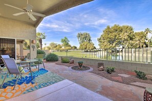 Une terrasse extérieure avec une table et des chaises, un tapis décoratif, un ventilateur de plafond et une vue sur un espace clos avec des arbres et une piscine.