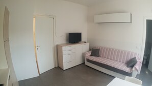 Ein kleiner, sauberer Raum mit weißer Tür, einem kleinen Schreibtisch, einem Fernseher auf einem Schrank, einem gestreiften Sofa und einer weißen Wand-Klimaanlage.