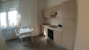 Eine kleine Küchenzeile mit Spüle, Herd und Mikrowelle. Ein weißer Tisch mit Stühlen und ein Fenster mit Jalousien.