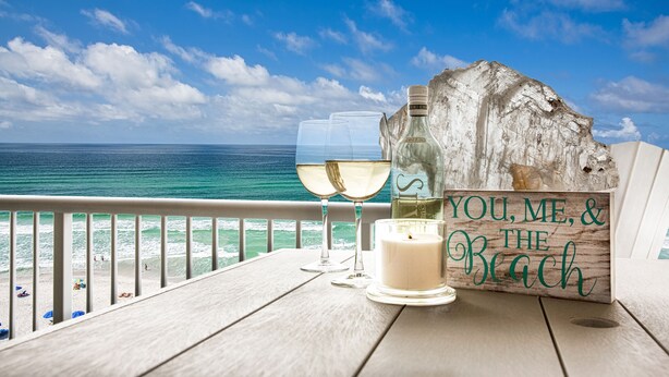 Ein Holztisch mit zwei Gläsern Weißwein, einer Kerze und einem Schild mit der Aufschrift 'YOU, ME, & THE BEACH'.