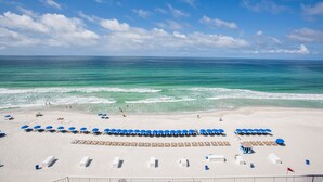 Ein Strand mit Reihen blauer Sonnenschirme und weißen Liegestühlen, ein klarer, blauer Himmel und das Meer im Hintergrund.