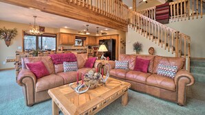 A spacious living room with a large brown leather sofa, a wooden coffee table, and a staircase with wooden railings. - Fire Pit + Views: Family Cabin on Table Rock Lake (Golden)