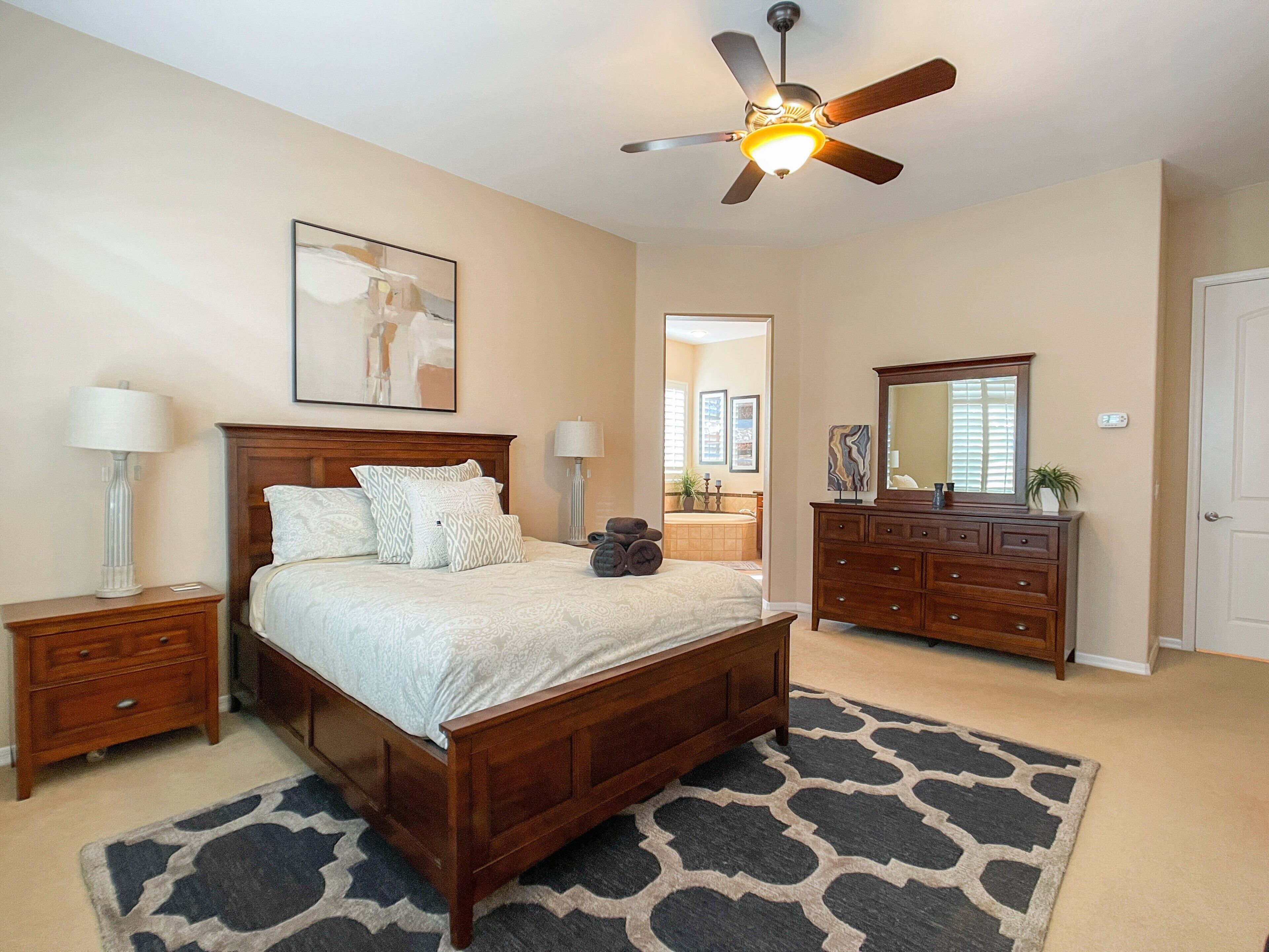 A bedroom with a wooden bed, a ceiling fan, a nightstand, a dresser, and a bathroom visible through an open door.