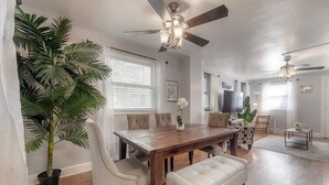 A dining area with a wooden table, chairs, and a bench. There are ceiling fans, a potted plant, and a television in the background.