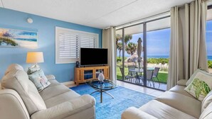 A hotel room with a TV, two sofas, a coffee table, and a large sliding glass door leading to a balcony with a view of the ocean.