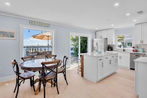 A modern kitchen with white cabinets, a dining area with a wooden table and chairs, and a view of the beach through a large window.