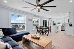 Un salon moderne comprenant un canapé, une table basse en bois, un coin repas avec des chaises, une cuisine avec des meubles blancs et un ventilateur de plafond.