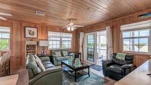 A cozy living room with wooden walls and ceiling, a glass coffee table, a sofa, a chair, and a dining table. - Sea Merc: A Charming Haven with Captivating Views in Hope Town (Hope Town)