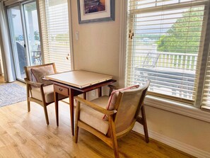 Una habitación con una mesa y sillas de madera, un cuadro en la pared y vistas a través de ventanas grandes con persianas.