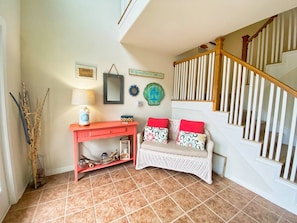 Un intérieur de maison comprenant un escalier, un bureau rouge, un canapé en osier blanc avec des coussins rouges et des œuvres d'art décoratives accrochées au mur.