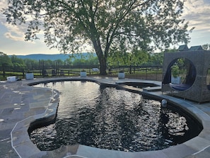 Una piscina de diseño curvo, rodeada de baldosas de piedra, un área de descanso con un ventanal circular y vistas a un árbol y un paisaje lejano.