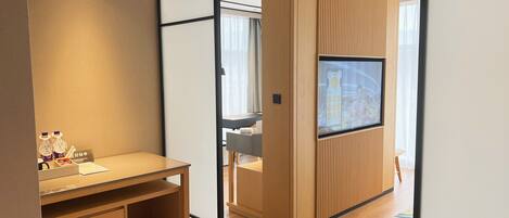 A modern hotel room with a wooden cabinet, a television mounted on a wooden panel wall, a small desk with a chair, and a round rug on the floor.