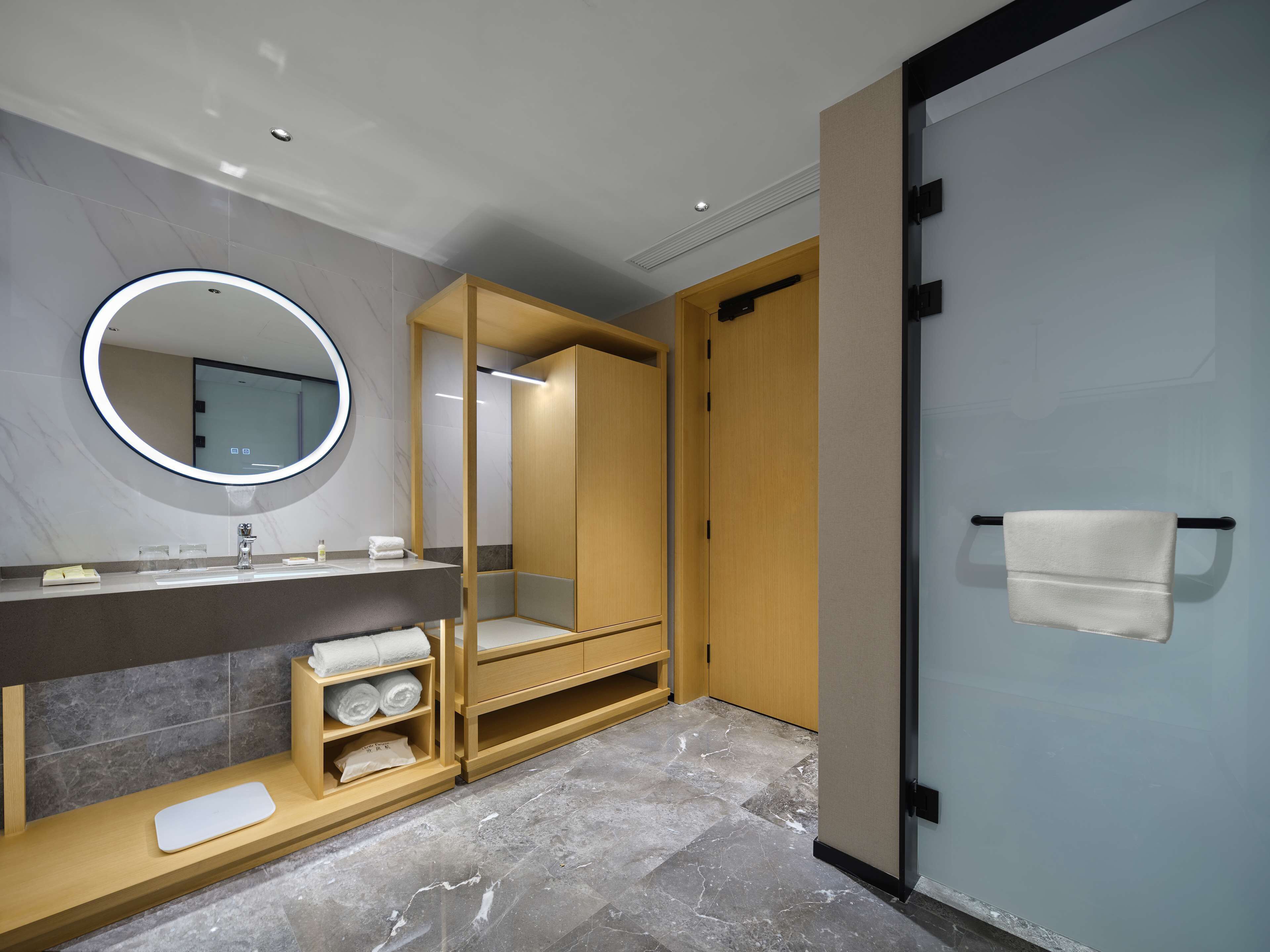 A modern bathroom with a marble floor, a round mirror with a light, a wooden vanity, and a towel rack.