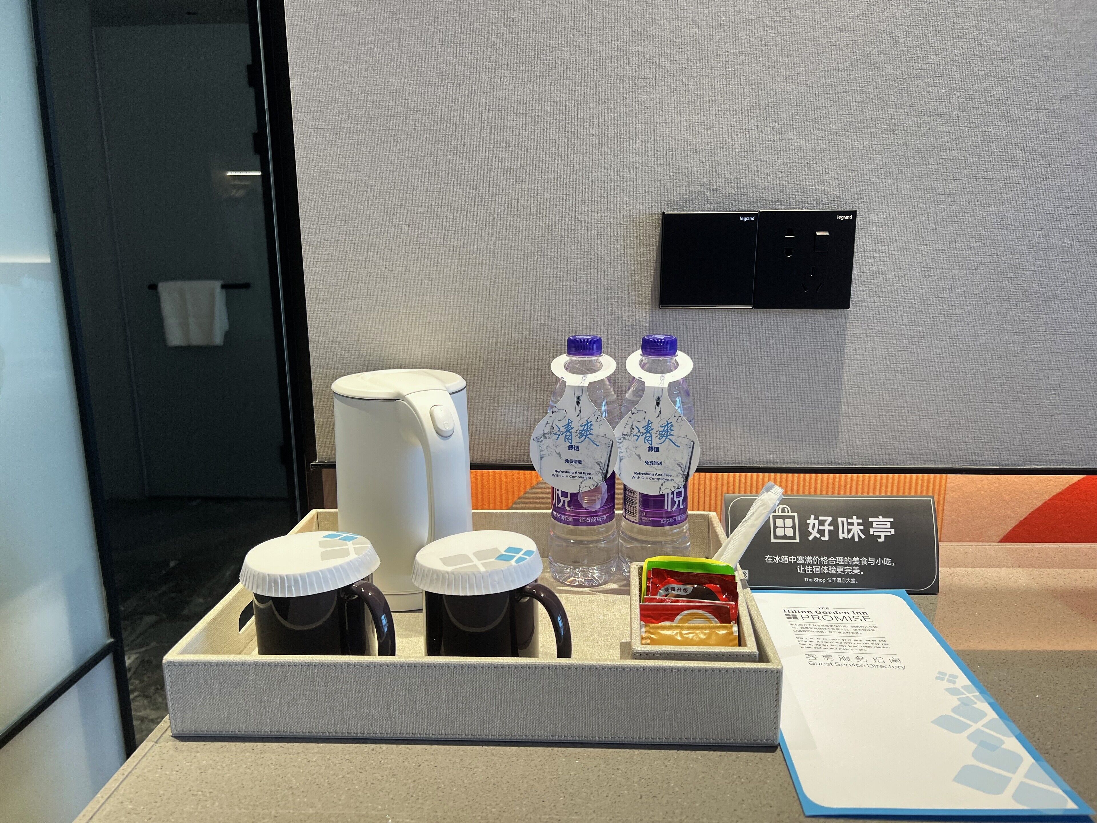 Zona de recepción de hotel con cafetera, dos botellas de agua, tetera y una pequeña caja de aperitivos.