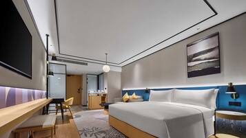 A modern hotel room with a large bed, a desk, a chair, and a small kitchenette.