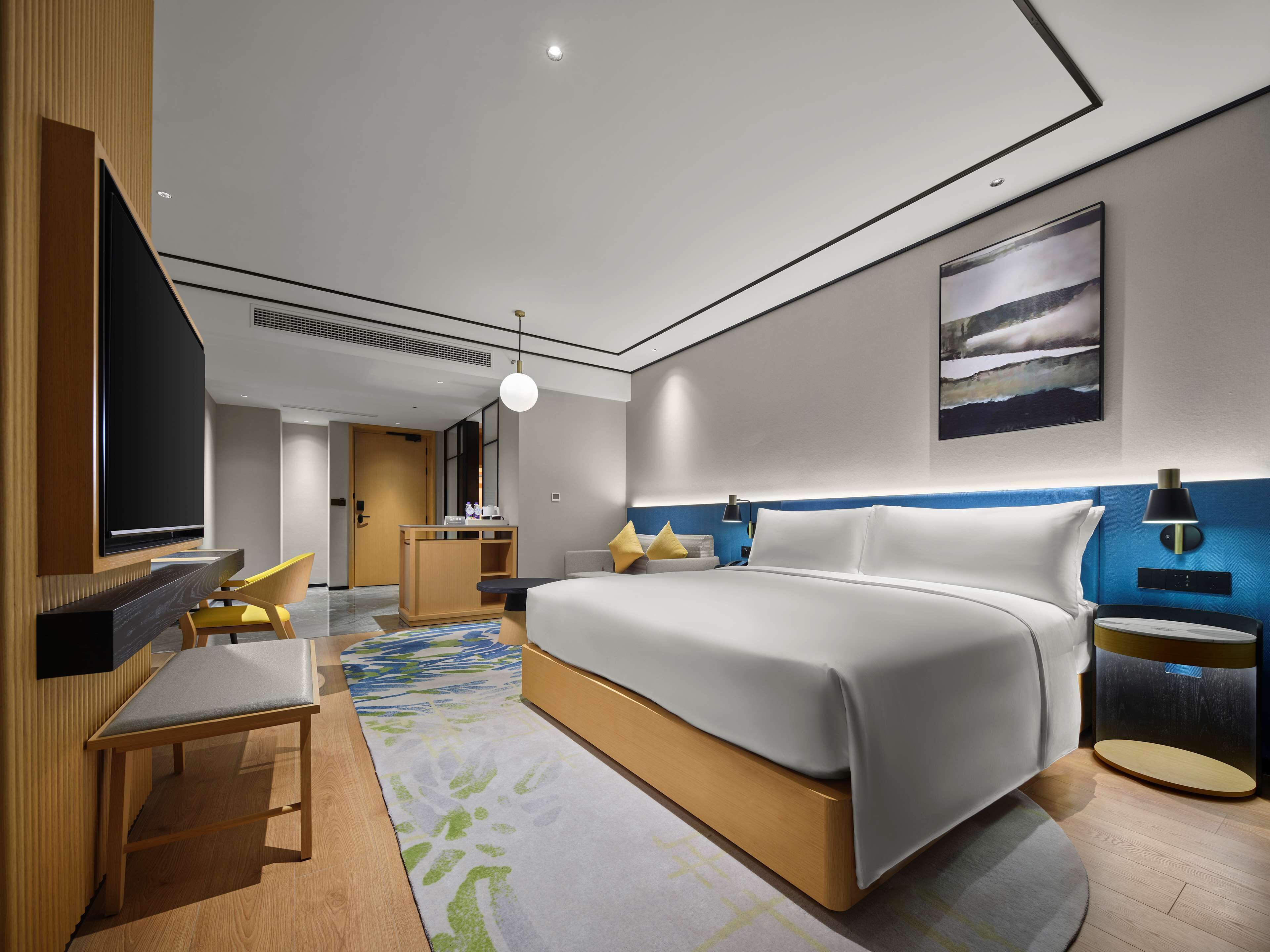Moderni hotellihuone, jossa on suuri sänky, työpöytä tuolilla, televisio ja taulu seinällä.