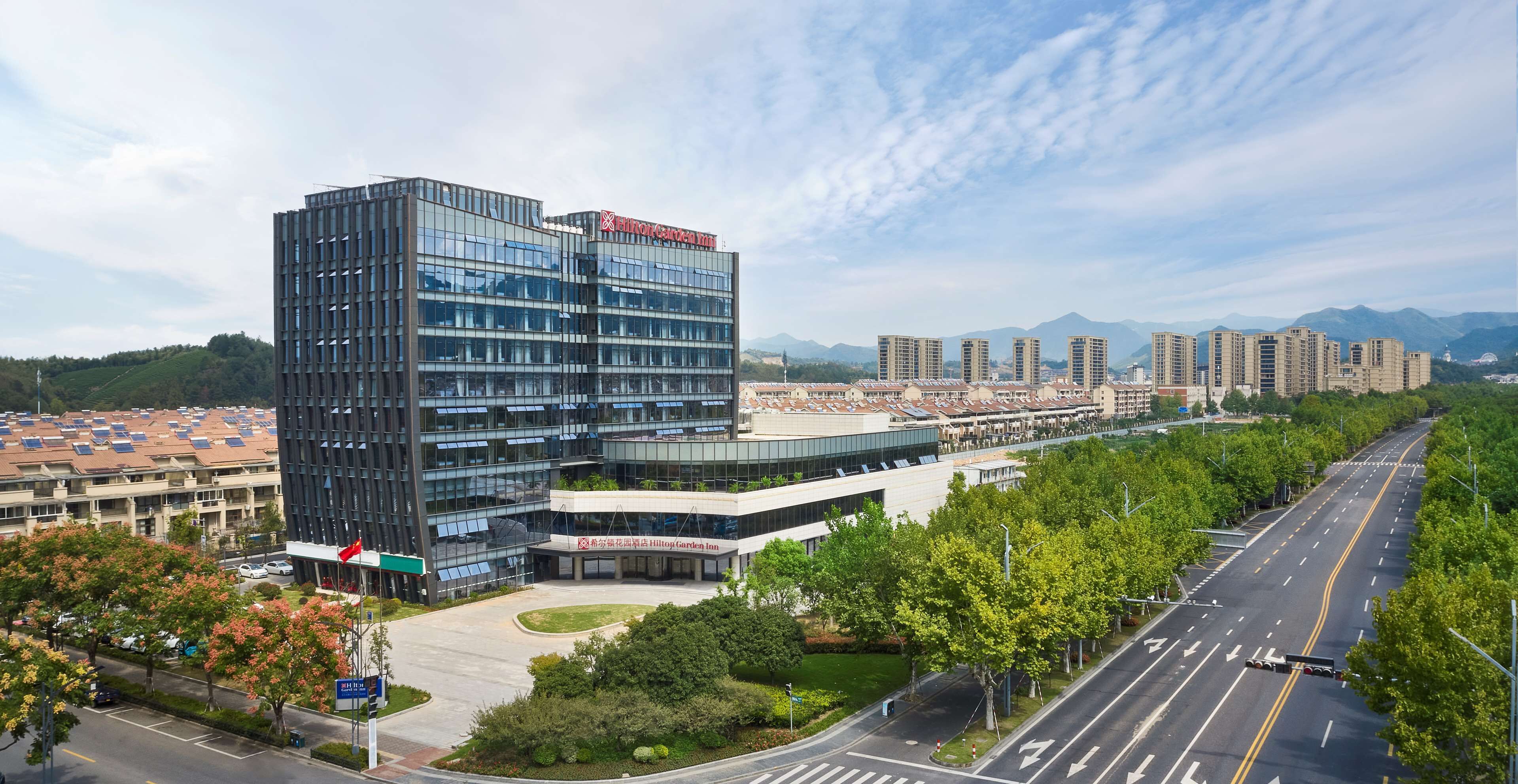 A modern hotel with a glass facade, a landscaped courtyard, and a view of surrounding buildings and mountains.