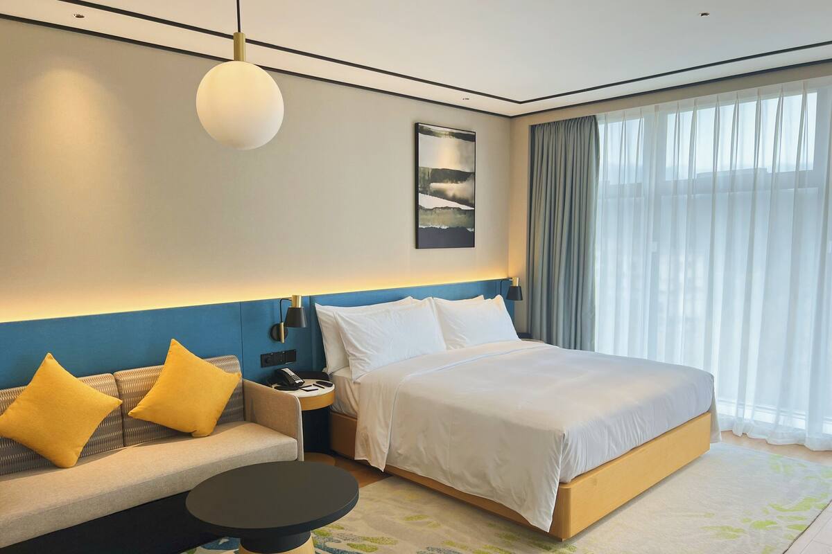 Habitación de hotel con una cama, un sofá con cojines amarillos, una mesita de noche con lámpara y un ventanal con cortinas.