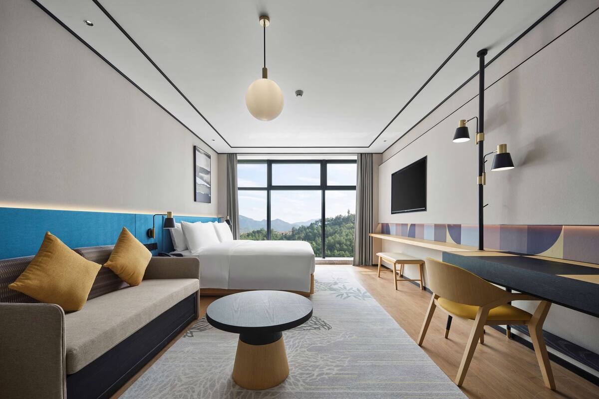 Una habitación de hotel moderna con cama, sofá, escritorio y ventanas amplias que dan a vistas de la montaña.