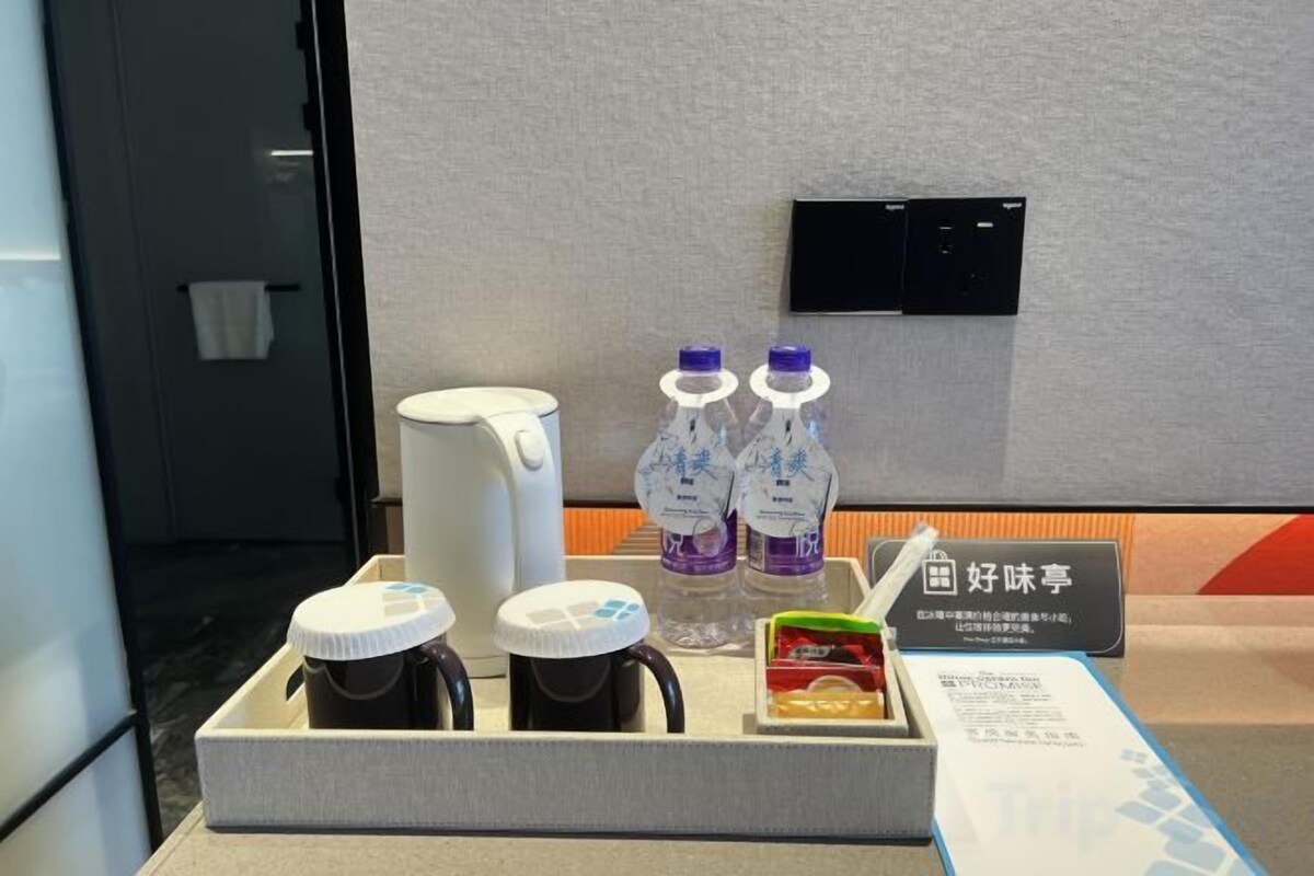 Zona de desayuno en una habitación de hotel, con una cafetera, dos botellas de agua, un jugo y un cartel con caracteres chinos.