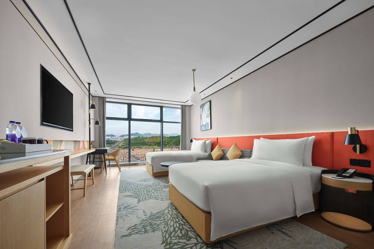 Habitación de hotel moderna con una cama grande, un escritorio y una silla. Hay un televisor montado en la pared y un ventanal que ofrece vistas de la ciudad.