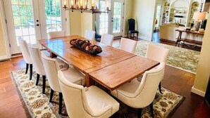 Un comedor con mesa de madera y sillas blancas tapizadas, una lámpara de araña y vista a la cocina al fondo.