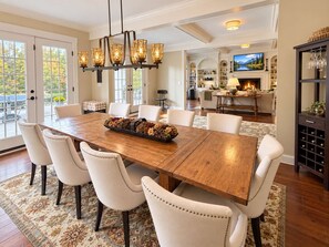A dining room with a wooden table and white upholstered chairs, a chandelier, and a fireplace in the background. - The Manor - Modern Farmhouse with Hot Tub (West Dover)