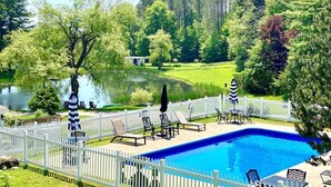 Una piscina con sillas de descanso y sombrillas, rodeada por una cerca blanca, con un lago y árboles al fondo.