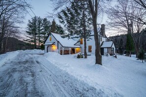Un paisaje nevado con una casa de dos pisos que tiene una chimenea, un porche con un brasero y una entrada de vehículos que conduce a la casa.