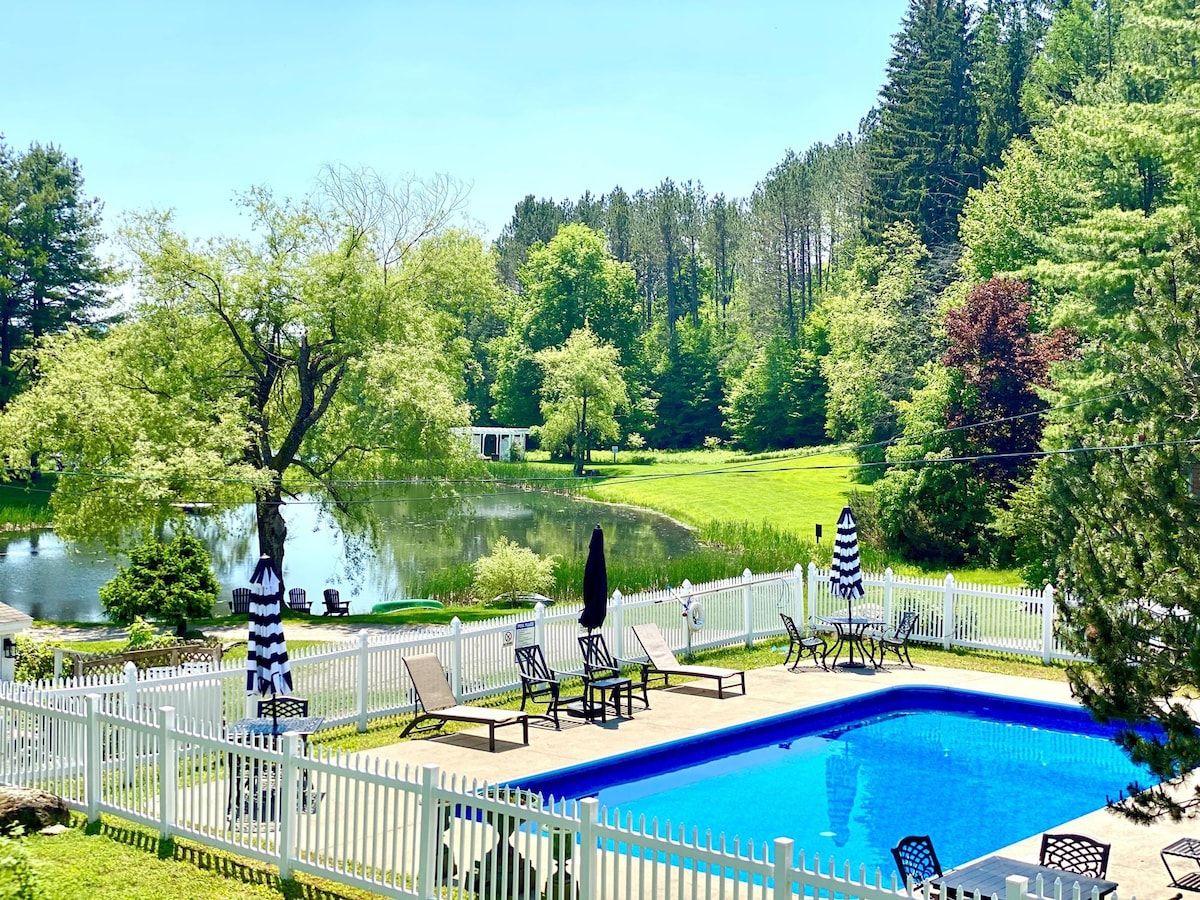 Una piscina con recinzione bianca, sdraio e vista su un lago e una foresta.