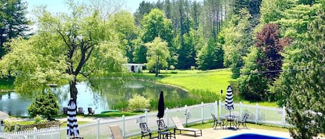 Una piscina con cerca blanca, sillas de descanso y vista a un lago y bosque.
