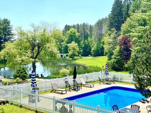 Una piscina con cerca blanca, sillas de descanso y vista a un lago y bosque.