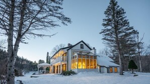Una casa de dos pisos con amplios ventanales y un patio cubierto, rodeada de nieve y árboles.