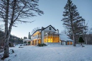 A two-story house with a glass facade, surrounded by snow-covered ground and trees. - The Manor - Modern Farmhouse with Hot Tub (West Dover)