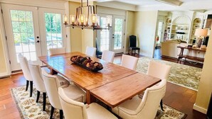 Un comedor con mesa de madera y sillas blancas tapizadas, una araña y vistas de la cocina y el comedor al fondo.