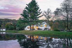 Une maison avec un grand arbre devant, un étang au premier plan et une colline boisée à l’arrière-plan.
