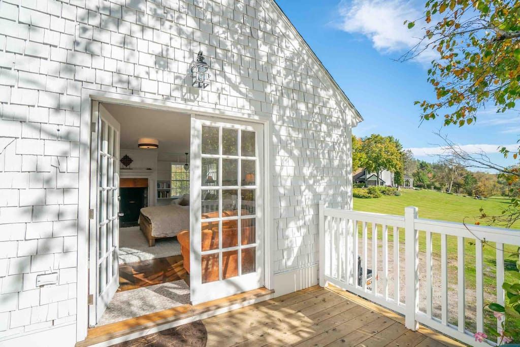 Una casa blanca con un porche, una puerta que da a una habitación con cama y ventana, y un patio cercado.