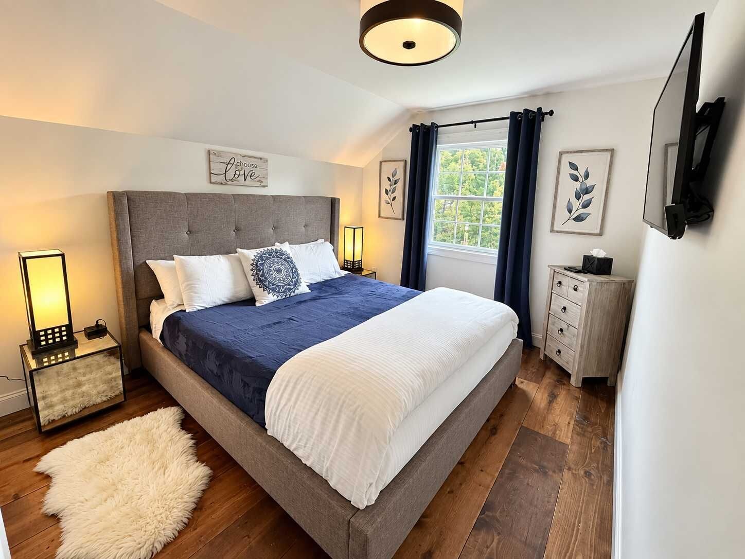 Un dormitorio ordenado con una cama grande, lámparas de noche, mesitas de noche y una alfombra mullida, además de una ventana con cortinas.