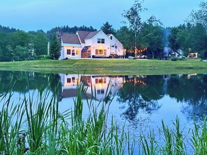 Une grande maison blanche, avec un étang devant, entourée d’arbres et d’herbe.