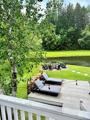 A deck with two lounge chairs, a fire pit, and a view of a pond surrounded by trees.